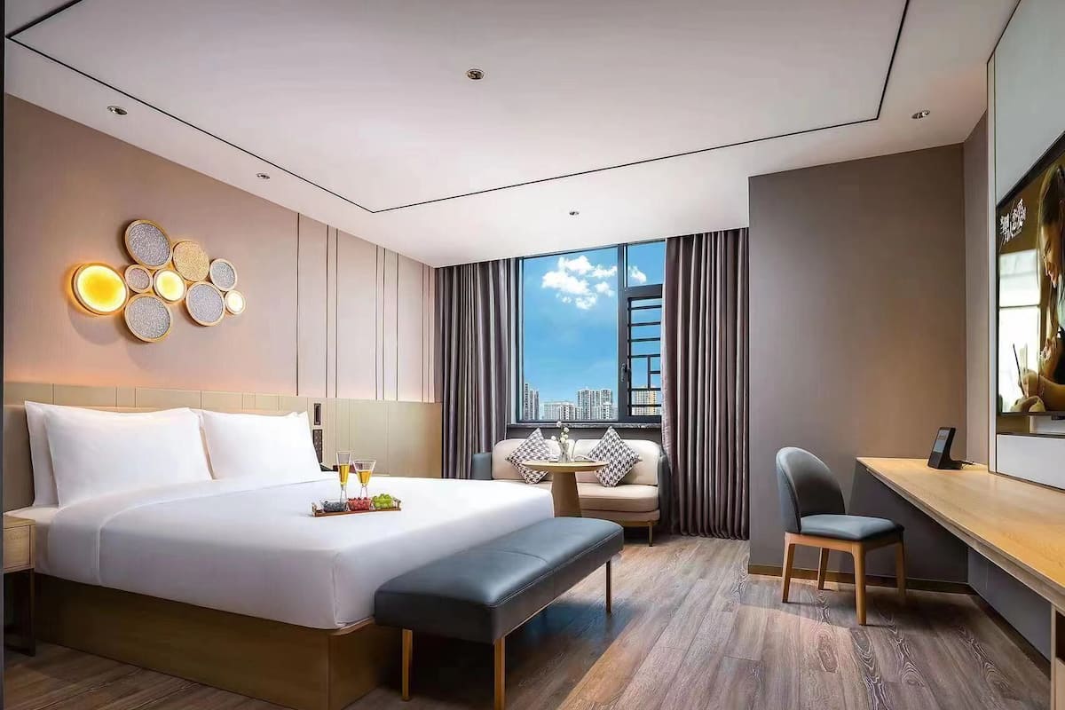 A modern hotel room with a large bed, a desk, a chair, and a view of the city skyline.