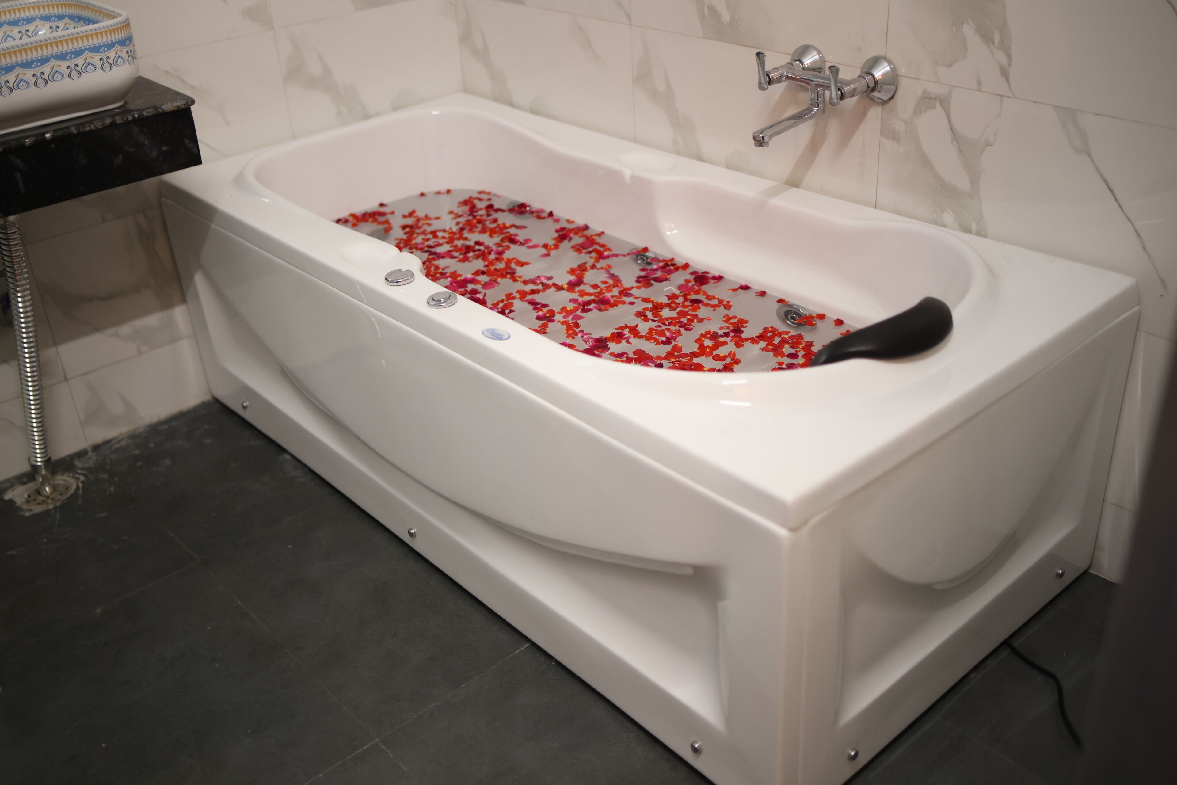 Una bañera blanca llena de agua y pétalos de rosa, con un tapete de baño negro y un recipiente blanco sobre una encimera negra.
