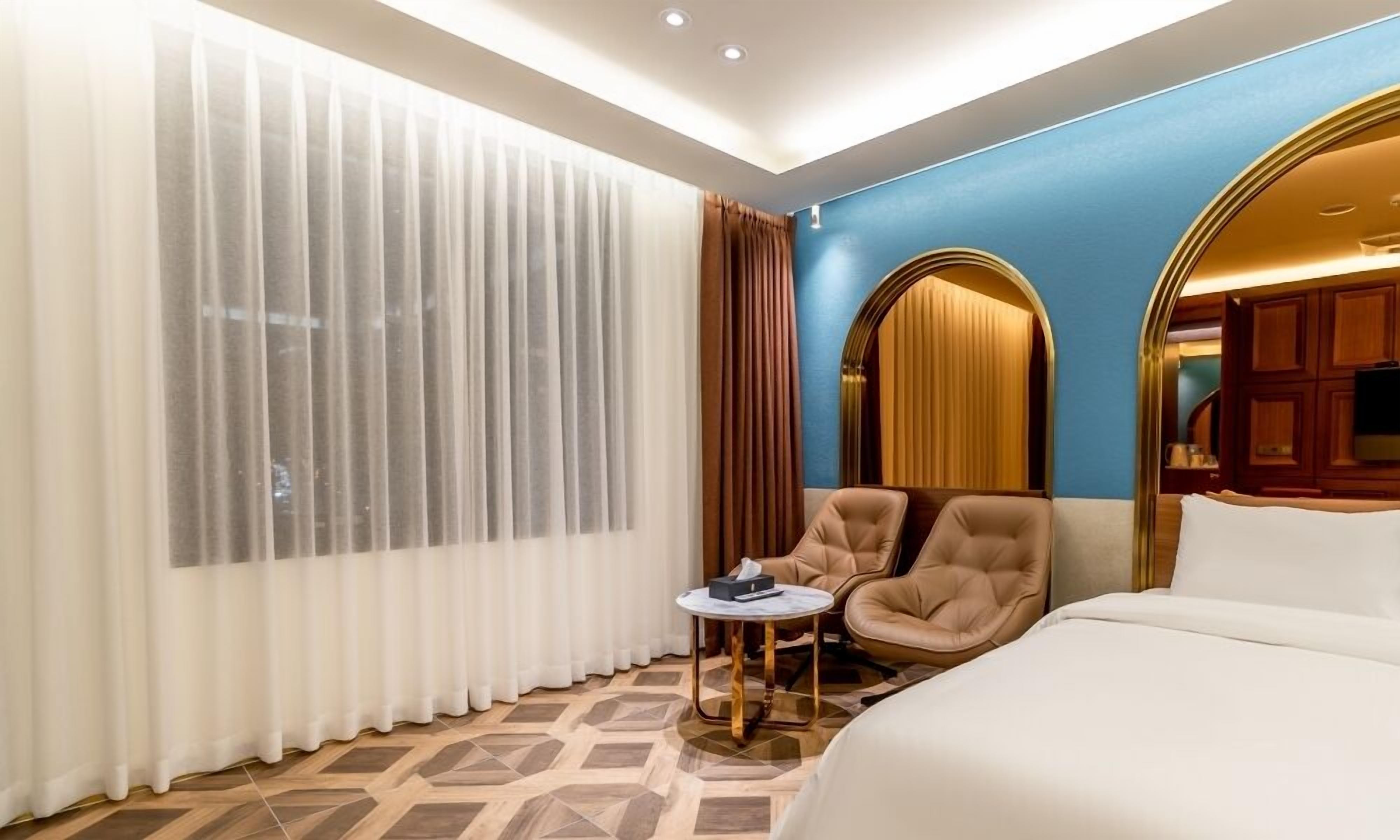 A hotel room with a bed, a chair, a small table, and a large window with curtains.