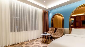 A hotel room with a bed, a chair, a small table, and a large window with curtains.