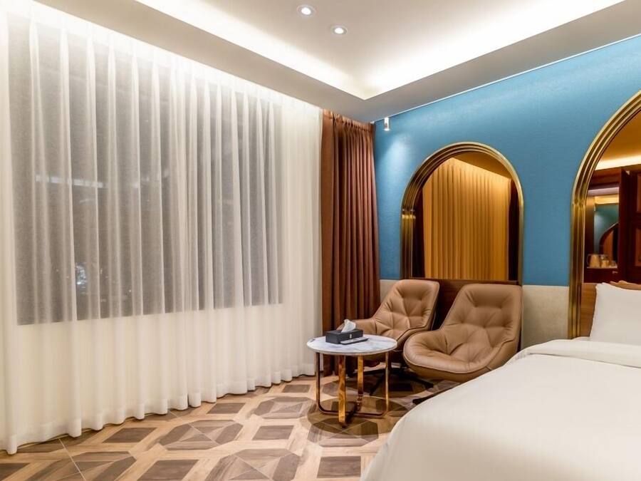 A hotel room with a bed, a chair, a small table, and a large window with curtains.