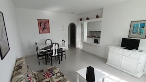 A living room with a sofa, dining table, chairs, a TV, and a kitchen area with cabinets and a microwave.