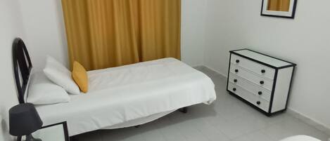 Un dormitorio con cama, mesita de noche con lámpara, cómoda y ventana con cortinas.
