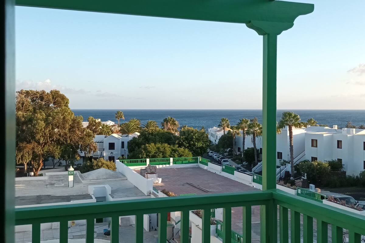 Un balcon donnant sur la mer, avec des bâtiments blancs et des balustrades vertes.