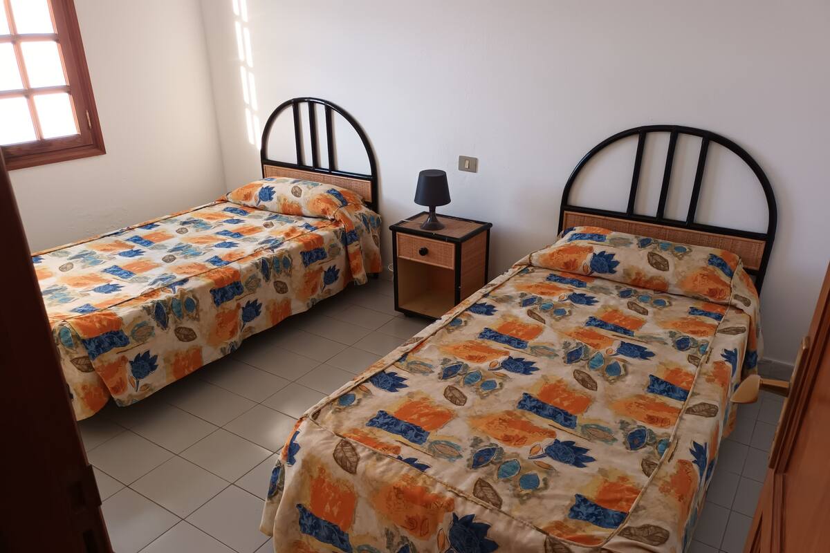 Une chambre avec deux lits simples, une table de chevet, une lampe et une fenêtre laissant entrer la lumière du soleil.