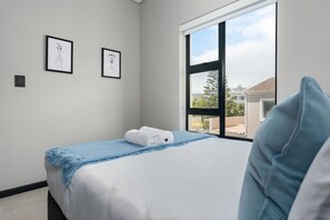 A neatly made bed with a blue blanket and white pillows, a window with a view of buildings, and two framed artworks on the wall.