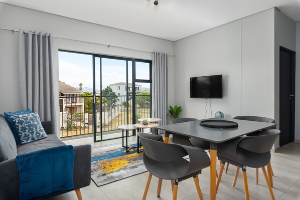 A modern living room with a sofa, a dining table with chairs, a TV, and a view of the outside.