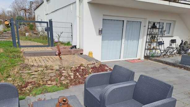 Un espace patio aménagé avec des meubles en osier, une petite table agrémentée d’un objet décoratif et un parterre de fleurs aux feuilles rouges.