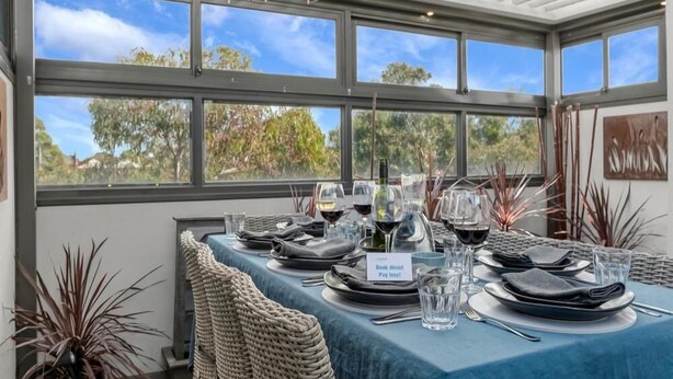 A dining area with a table set for four, a 'Book direct Pay less!' sign, and a view of trees and sky through large windows.