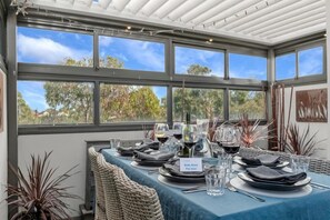 Uma área de refeições com uma mesa posta para quatro pessoas, um aviso 'Reserve direto Pague menos!' e vista para árvores e céu através de grandes janelas.