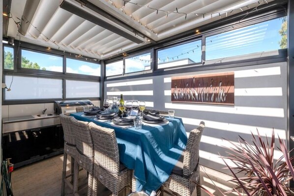 Uma área de refeições ao ar livre com cadeiras de vime e uma mesa posta para uma refeição, com toalha de mesa azul e uma garrafa de vinho.