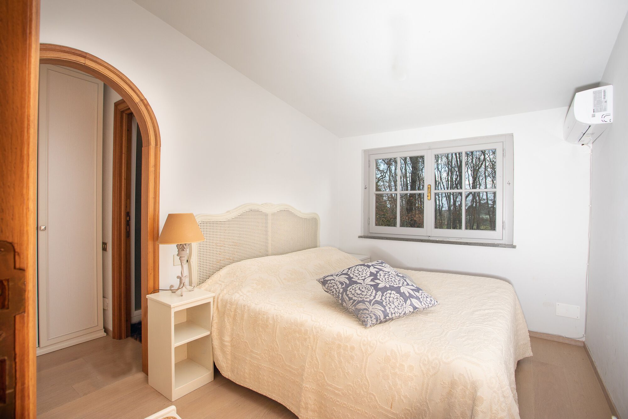 A bedroom with a bed, a nightstand with a lamp, a window with a view of trees, and a door.