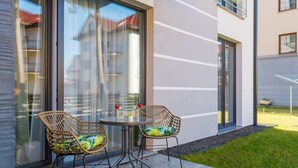 A modern outdoor seating area with wicker chairs and a glass table, adjacent to a building with large windows and a grassy lawn.