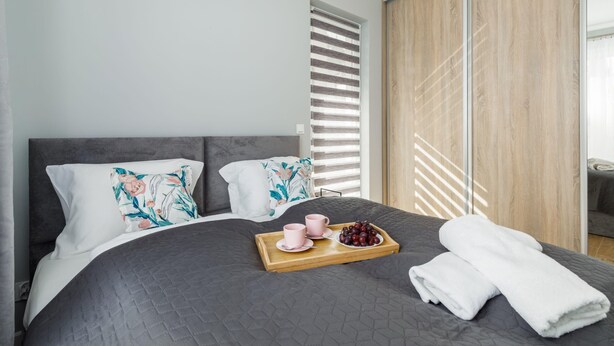 A modern bedroom with a large bed, a wooden side table with a tray of fruit and cups, and a view of a wooden sliding door.