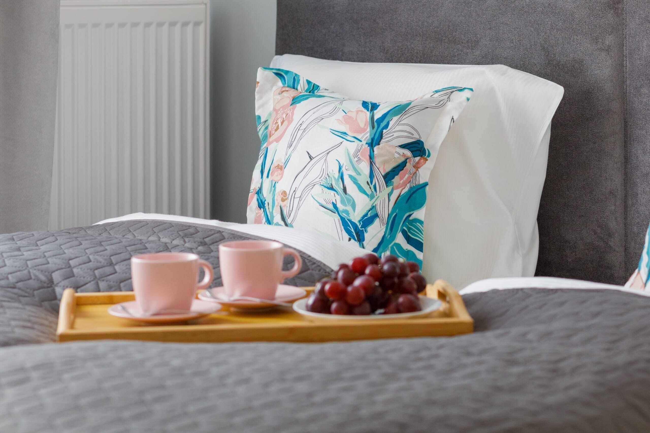 Un lit bien fait, recouvert d’une couverture grise, avec un oreiller blanc orné d’un motif floral et un plateau sur lequel on trouve une tasse rose, une soucoupe et des raisins.