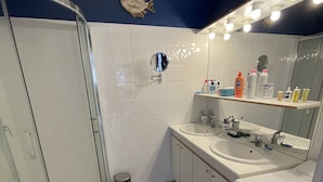 Une salle de bain avec une douche à parois de verre, des murs recouverts de carreaux blancs, un lavabo avec un miroir et un tapis de bain noir.