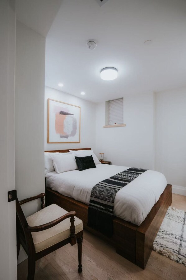 A bedroom with a bed, a chair, a bedside table, a lamp, and a framed picture on the wall.