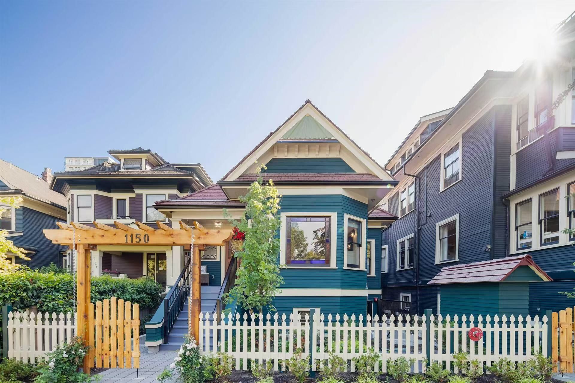 A row of townhouses with a wooden pergola labeled "1150" in front, a white picket fence, and a clear blue sky.