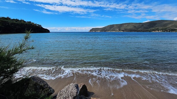 A beach with clear water, rocky foreground, and hills in the background.