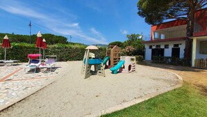 A playground with slides and climbing structures, surrounded by lounge chairs and umbrellas, with a building in the background.