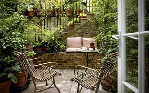 Une terrasse de jardin accueillante, avec un mur en pierre, un escalier en bois et des plantes en pot. On y trouve deux fauteuils en osier et une petite table avec des tasses et des soucoupes.