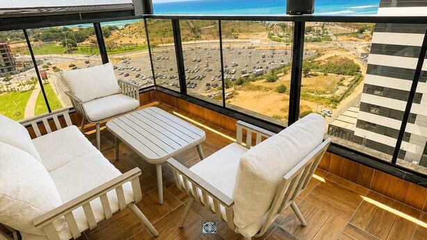 Un balcone con pavimento in legno, sedie imbottite bianche, un tavolino e vista sull'oceano e sulla città.
