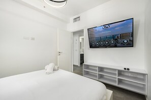 A modern hotel room with a large flat-screen TV mounted on the wall, a neatly made bed, a white console table, and a door leading to a bathroom. - Luxury 4BR Tower w/ Panoramic Sea View | Mamad (Bat Yam)