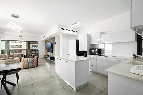 A modern kitchen with white cabinetry, a central island, and a dining area with a table and chairs. - Luxury 4BR Tower w/ Panoramic Sea View | Mamad (Bat Yam)