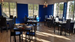 Un comedor con varias mesas y sillas, amplias ventanas, paredes azules y suelo de madera.