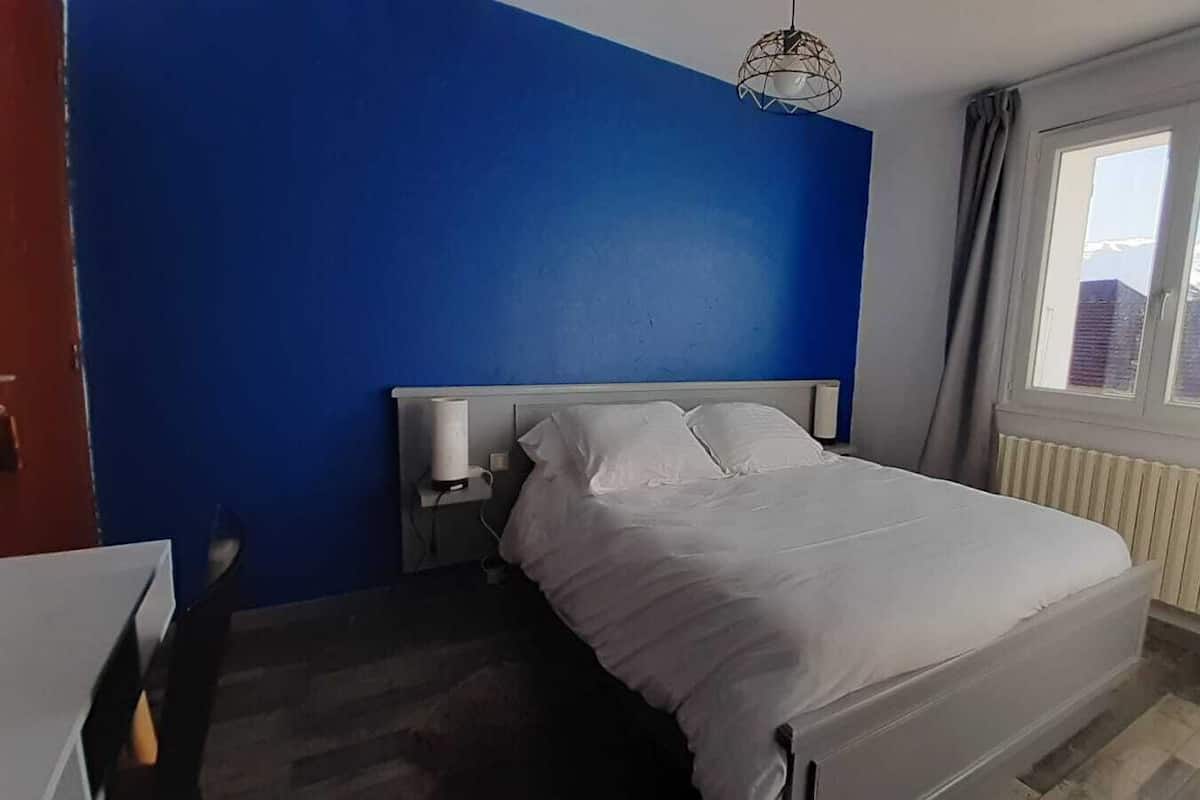 A bedroom with a bed, a bedside table, a lamp, a window, and a view of the outside.
