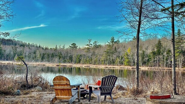 A lakeside setting with two Adirondack chairs facing a calm lake, surrounded by trees and a clear sky.