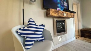 A modern living room with a white chair, a blue and white striped pillow, a mounted television displaying streaming services, and a fireplace with a rustic mantel.