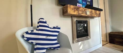 A modern living room with a white chair, a blue and white striped pillow, a mounted television displaying streaming services, and a fireplace with a rustic mantel.