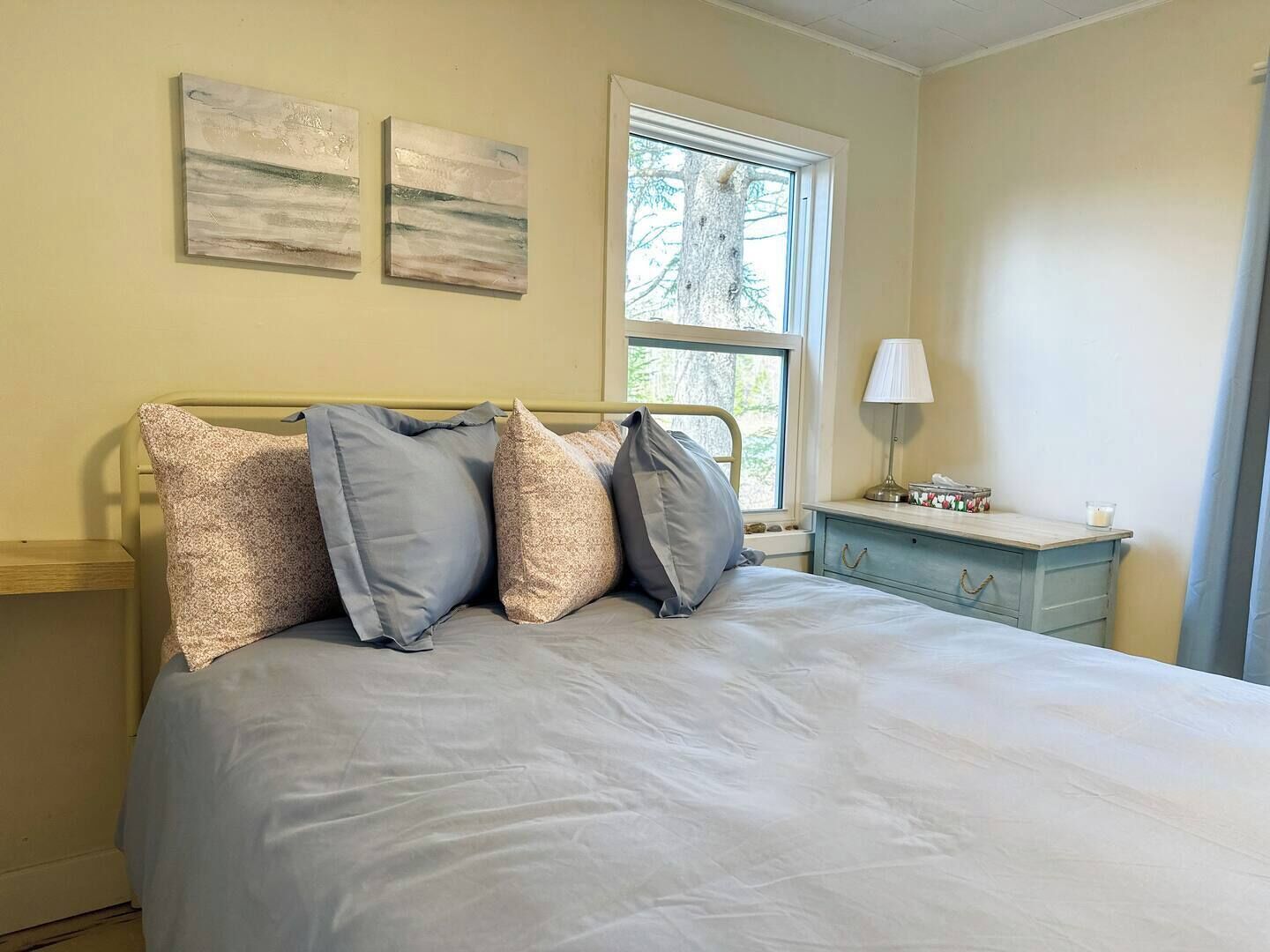 Un dormitorio con cama, almohadas, una mesita de noche, una lámpara y dos cuadros en la pared.