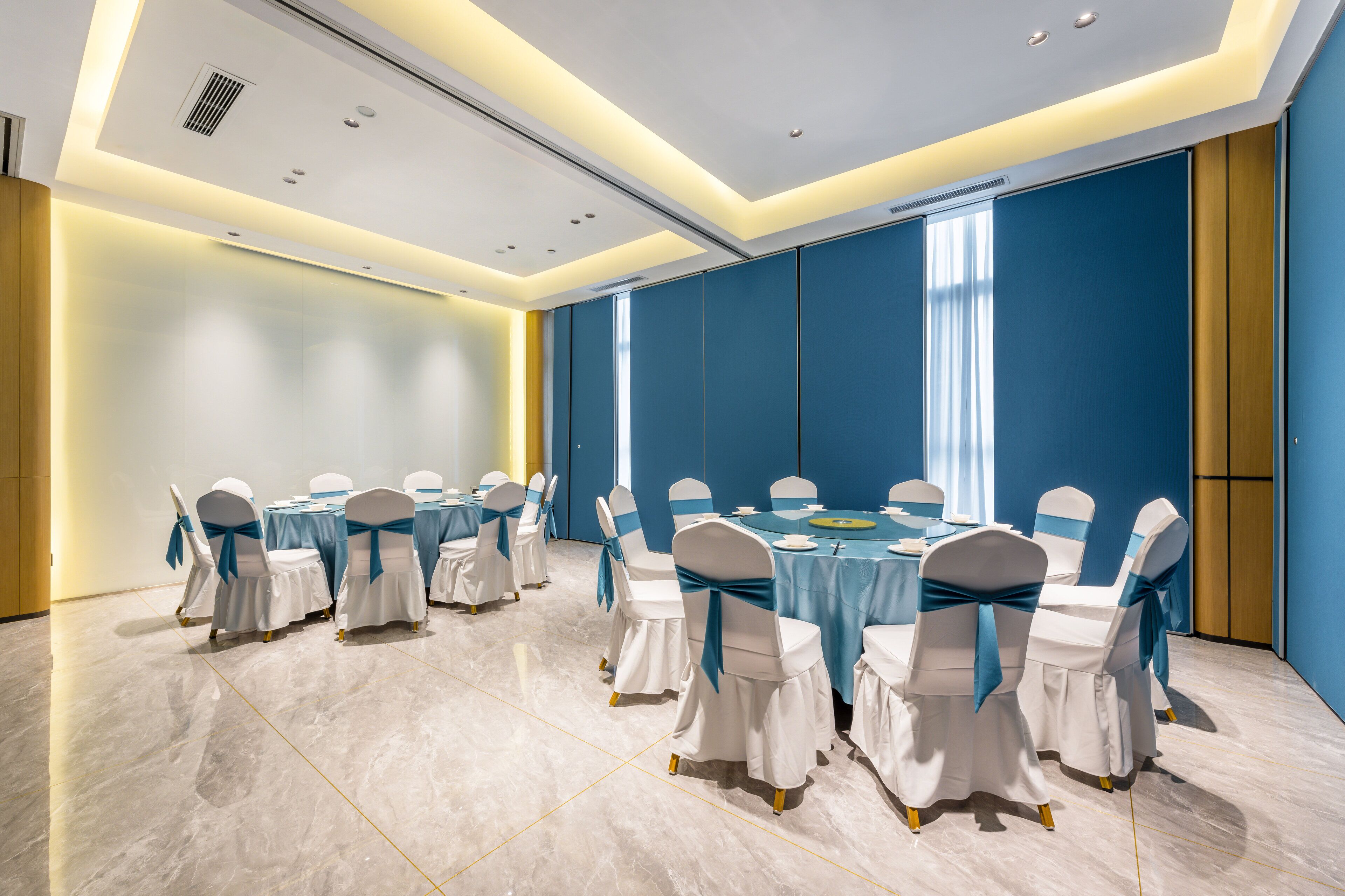Un salón de conferencias con mesas redondas, sillas blancas con cintas azules y una pared de acento en tono azul.