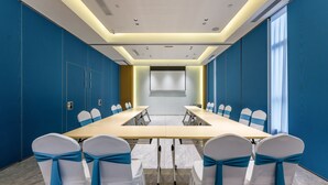 Une salle de conférence avec une longue table, des chaises blanches ornées de rubans bleus et un grand écran à l’avant.