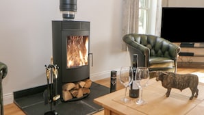 A cozy living room with a wood-burning stove, a green leather sofa, a wooden coffee table with wine glasses and a decorative sheep figurine, and a television in the background. - Dove Meadows House (Buxton)