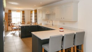A modern kitchen with dark cabinetry, a large island, and a dining area with chairs and a table. - Dove Meadows House (Buxton)