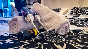 Een bed met een zwart-wit bloemenbedekking, een opgerolde witte handdoek, een fles wijn en een paar brillen.