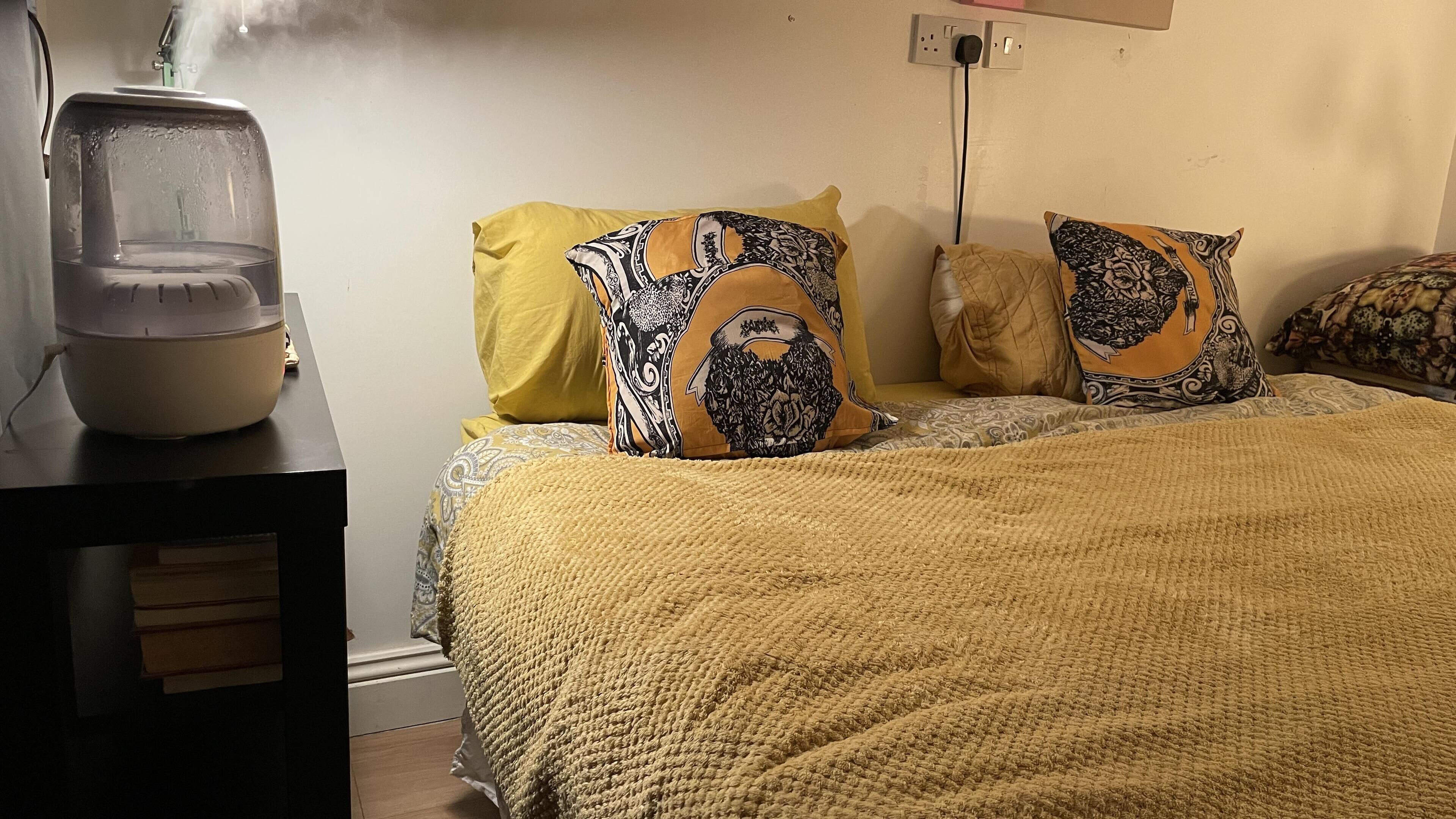 A bed with a yellow blanket and patterned pillows, a bedside table with a water purifier, and a wall socket.