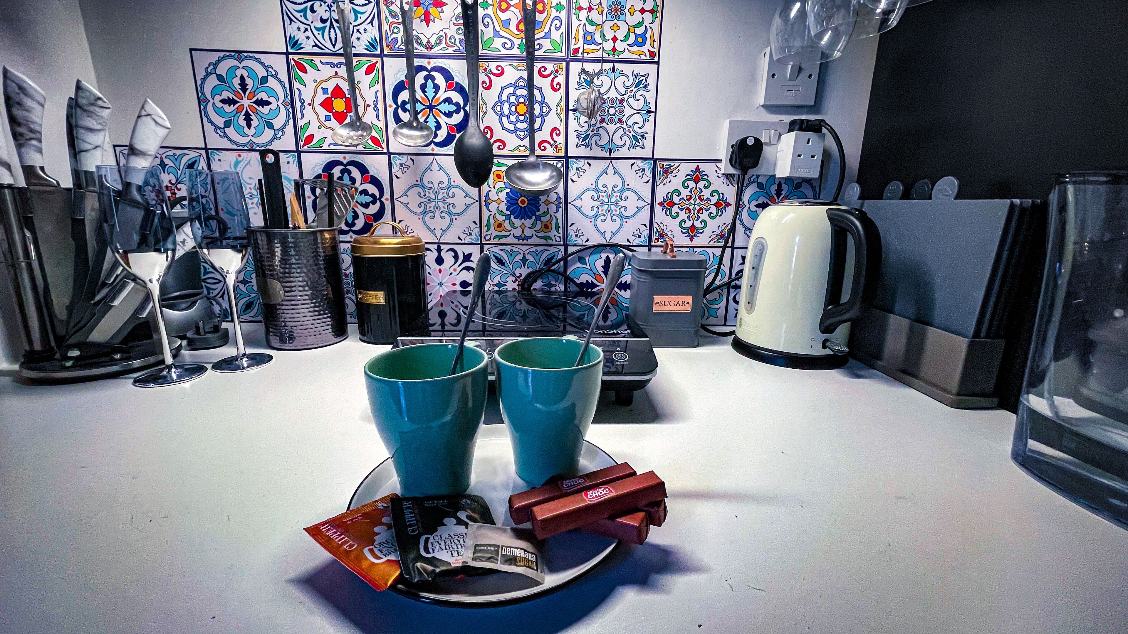 A kitchen with a tiled backsplash, a coffee maker, a kettle, and two blue cups on the counter.