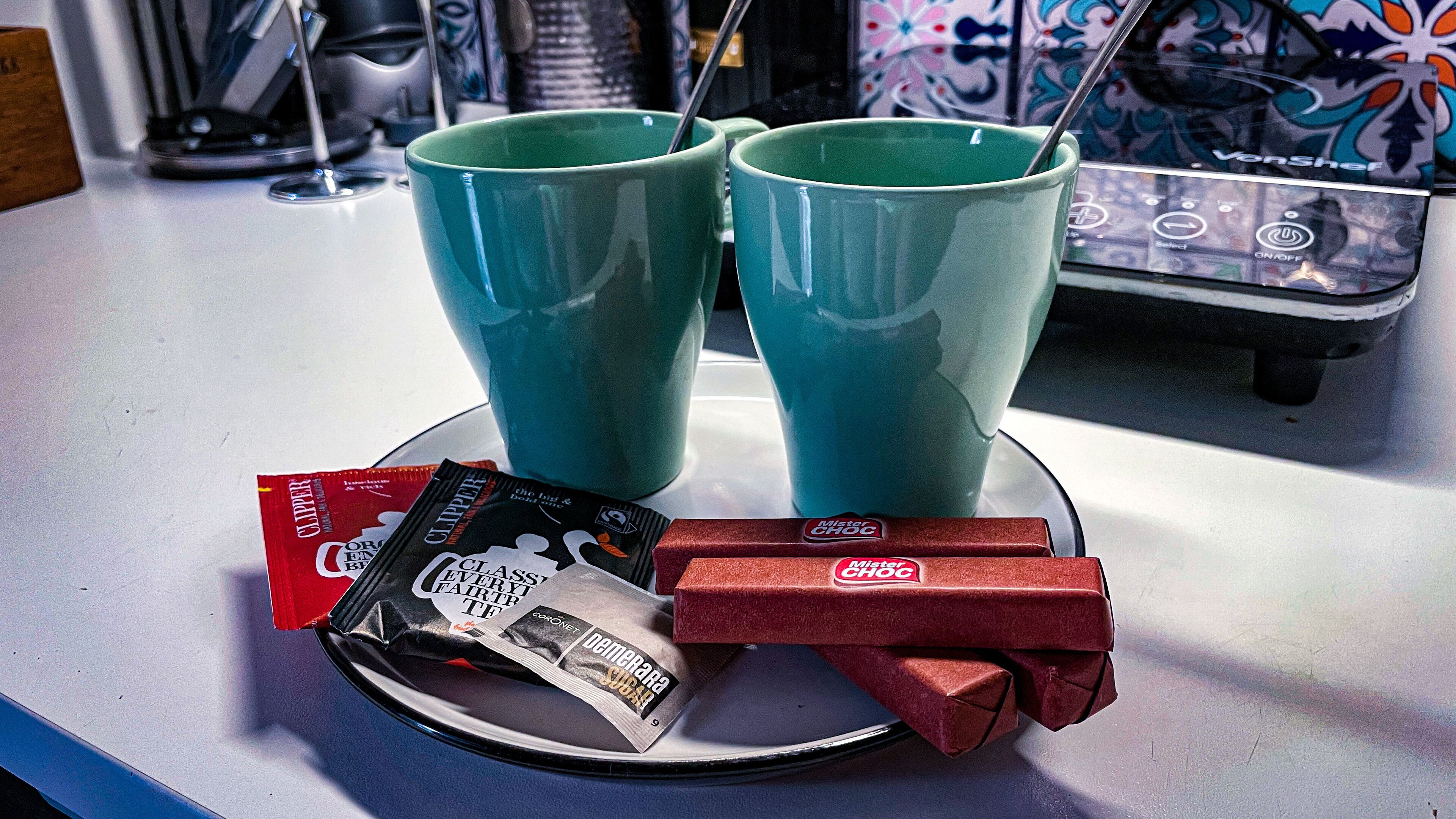 Un piano di lavoro in cucina con due tazze color petrolio, un piatto con barrette di cioccolato e una bustina di zucchero.
