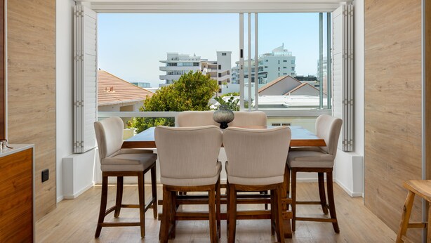 A dining area with a table and chairs, large windows, and a view of buildings and trees. - Seacliffe 307 (Cape Town)