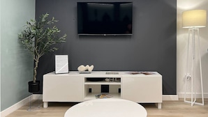 A modern living room with a grey wall, a mounted TV, a white entertainment unit, a potted plant, and a floor lamp. - Underfloor heating  & Free Parking in the city center (Debrecen)