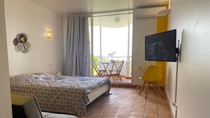 A bedroom with a bed, a chair, a TV, and a balcony with a table and chairs. - Studio "le pipiri" swimming pool sea view secure residence (La Trinité)