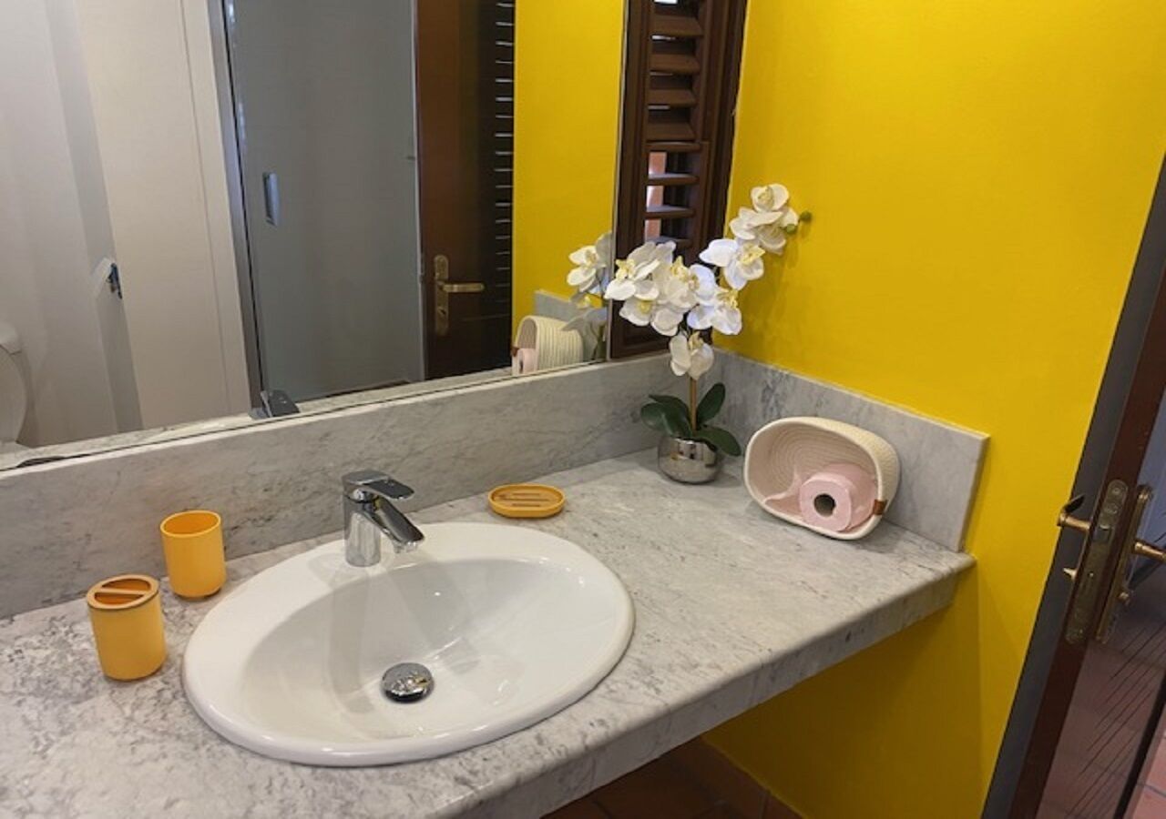 Ein Badezimmer mit einem weißen Waschbecken, gelben Wänden, einem Spiegel und einer Topforchidee.