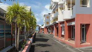 A street lined with multi-story buildings, some with balconies, and palm trees along the sidewalk. - Studio "le pipiri" swimming pool sea view secure residence (La Trinité)