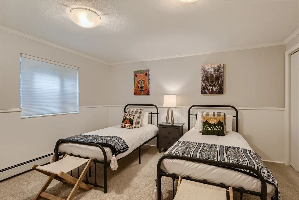 A bedroom with two single beds, a nightstand, a small table, and a window with blinds.