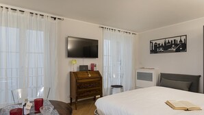 A hotel room with a bed, a TV, a wooden dresser, a desk with a lamp, and a cityscape wall art. - Superba Studio by Wonderful Italy (Genoa)