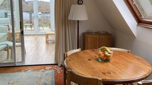 A dining area with a round wooden table, chairs, and a fruit bowl. A lamp and a cabinet are also visible.